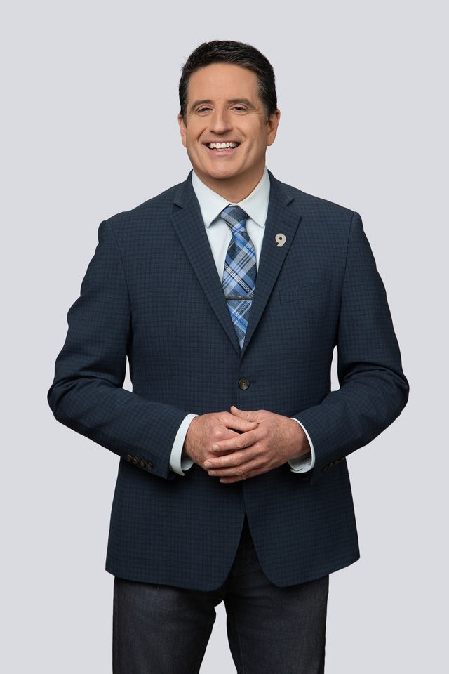 A man in a dark blue blazer, light blue shirt, and plaid tie stands smiling with hands clasped in front of a plain light gray background.