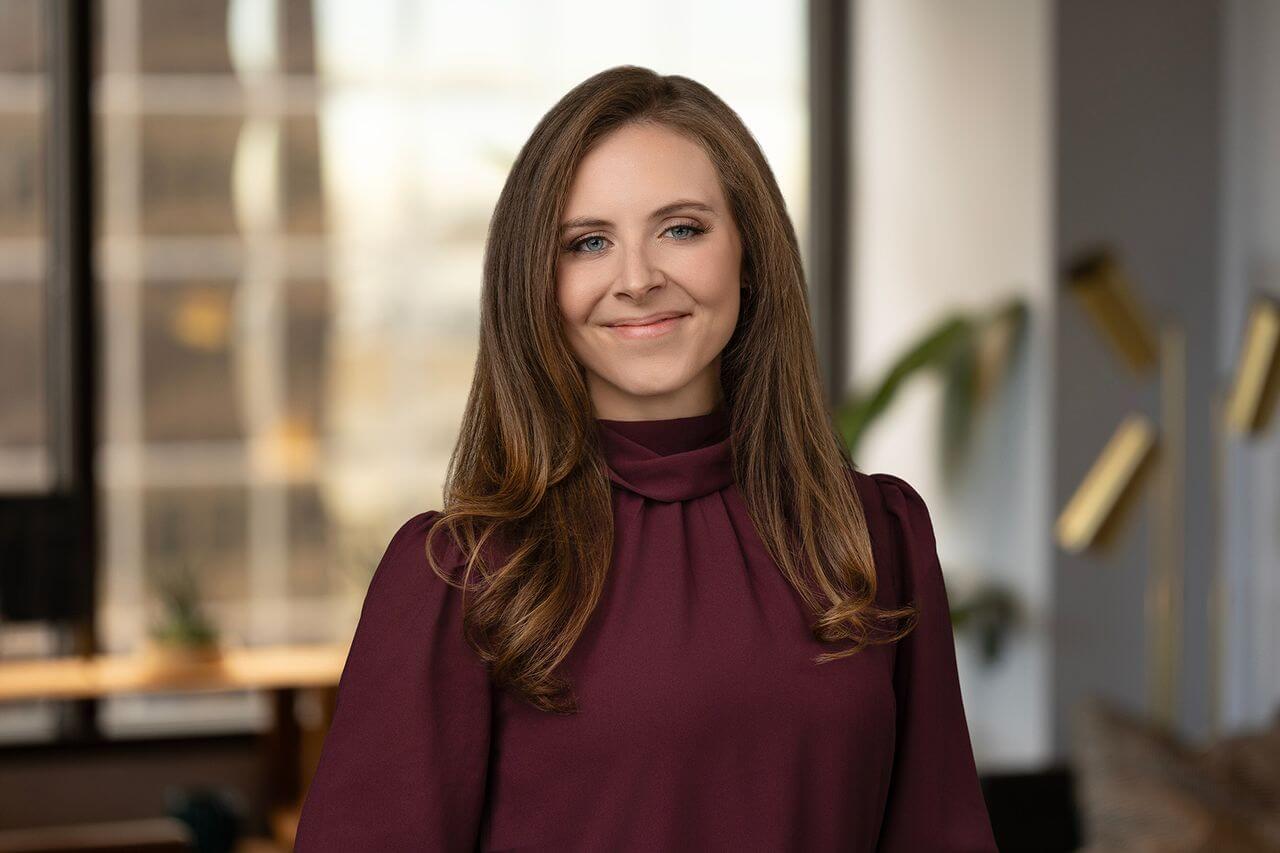 A woman with long brown hair, wearing a maroon blouse, stands indoors in front of large windows and modern office decor, smiling at the camera.