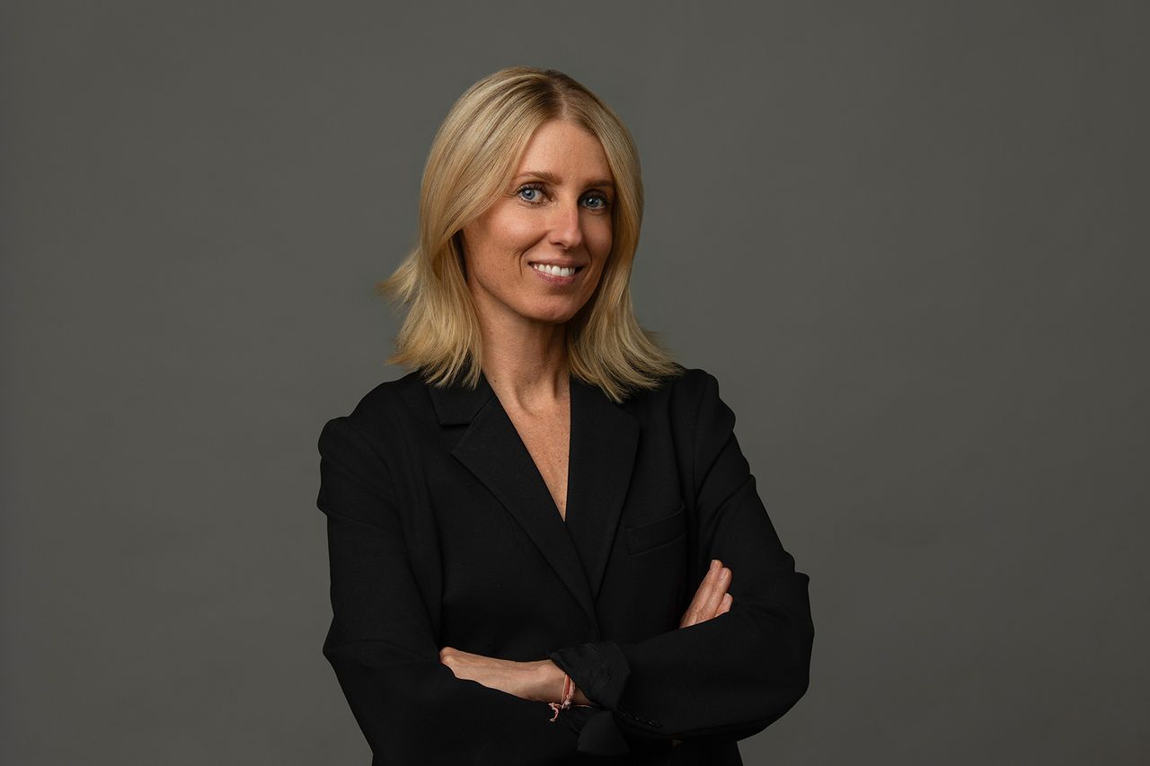 A woman with shoulder-length blonde hair wearing a black blazer stands with arms crossed against a plain gray background.