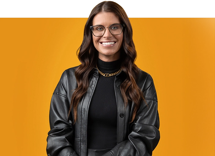 A woman with long brown hair, wearing glasses, a black leather jacket, and a gold chain necklace, stands smiling in front of an orange background.