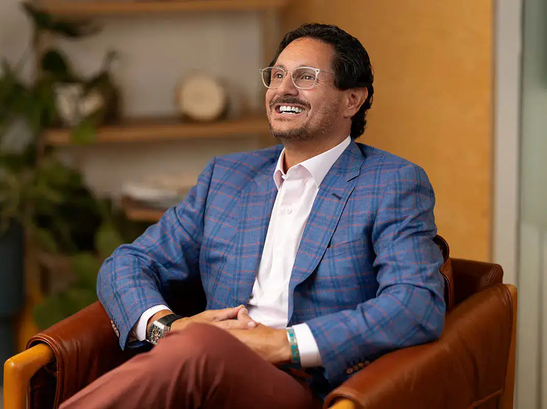 A man with glasses and a blue plaid jacket sits in a brown leather chair, smiling, with shelves and a plant in the background.
