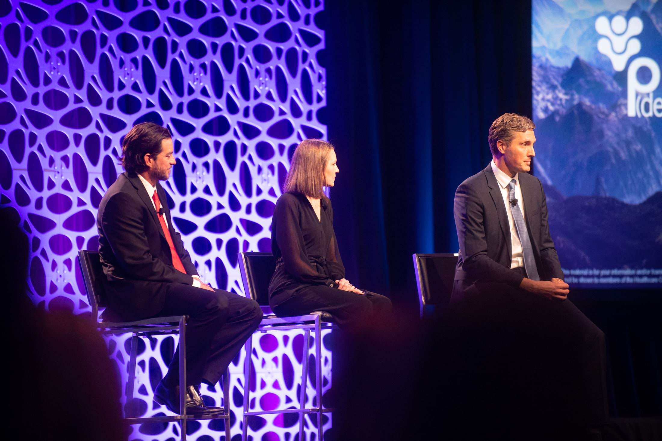 three people sitting on stage in front of a large screen.