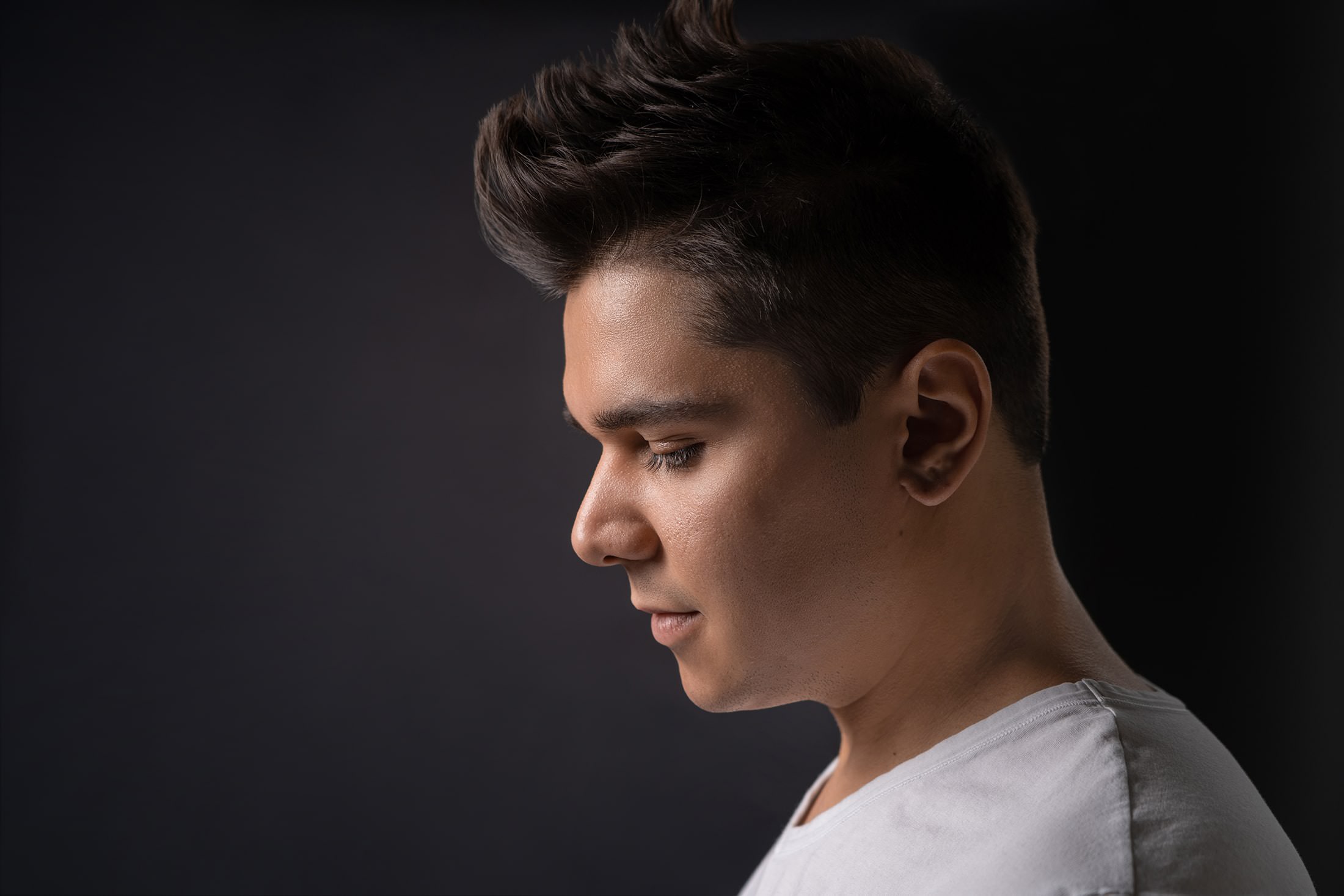 A young man in a white shirt illuminated dramatically against a jet black background, emphasizing contrast and conveying a sense of mystery.