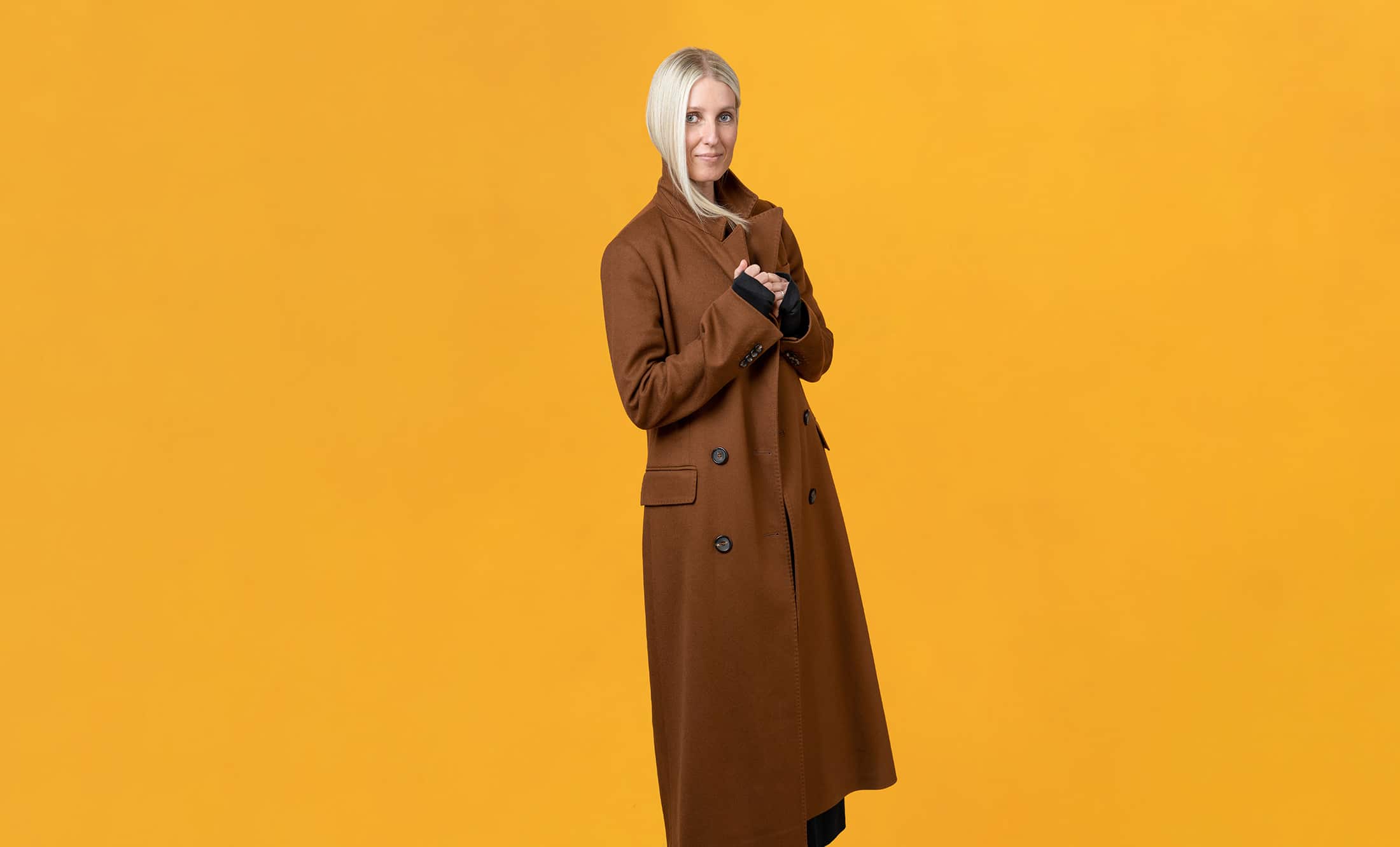 a woman in a brown coat posing on a yellow background.