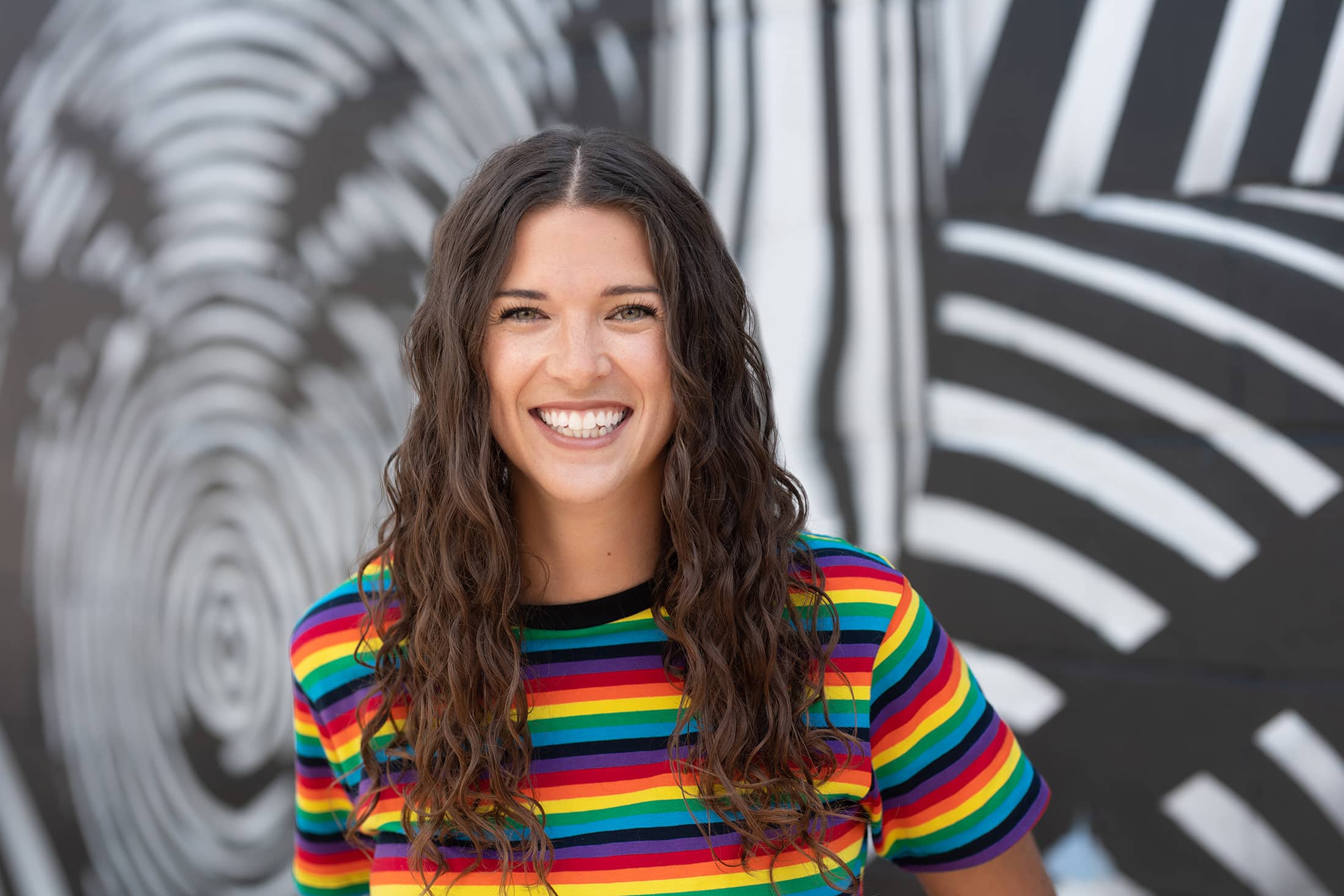 a woman in a rainbow striped shirt smiling in front of a wall.