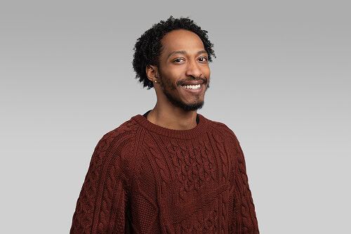 A man with short curly hair and a beard, wearing a brown cable-knit sweater, smiles at the camera against a plain gray background.