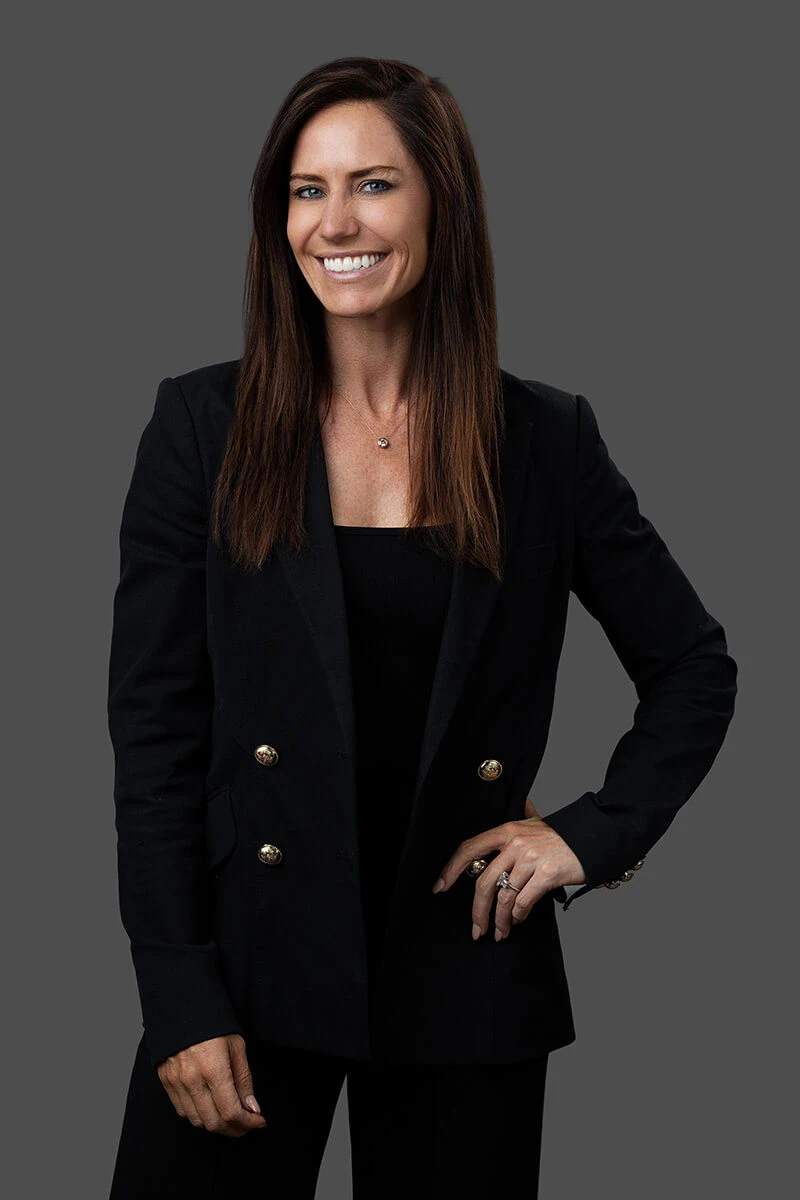 A woman with long brown hair wearing a black blazer and black top stands against a gray background, smiling with one hand on her hip.