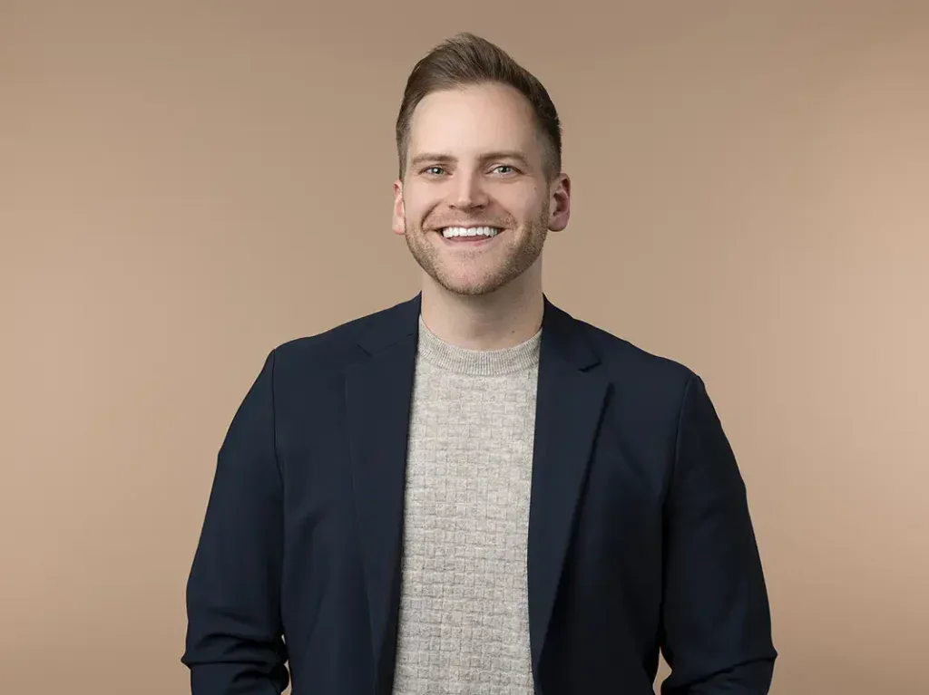 A man wearing a navy blazer and light gray sweater stands smiling against a plain beige background.