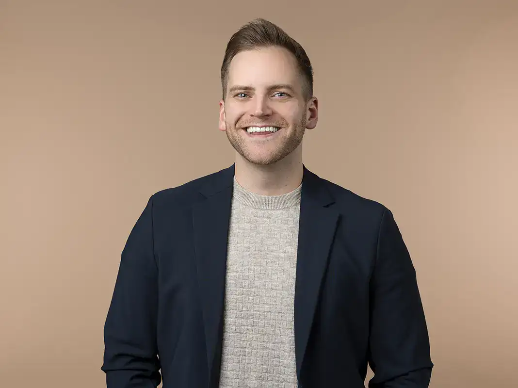 A man wearing a navy blazer and light gray sweater stands smiling against a plain beige background.