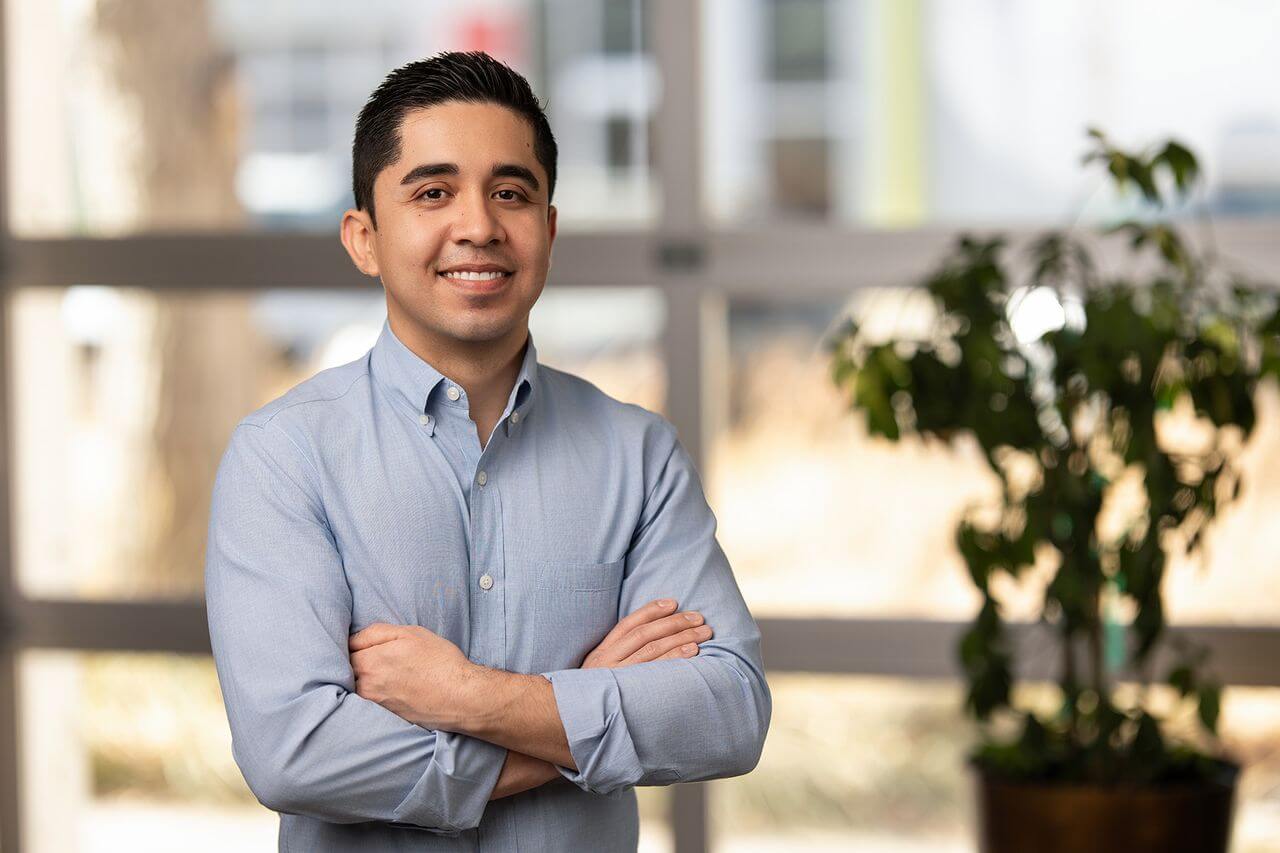 A man in a light blue button-up shirt stands indoors with his arms crossed, smiling. A potted plant and large windows are visible in the background.