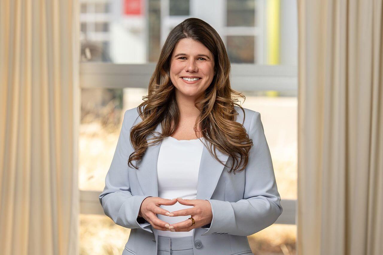 Woman in a light gray suit and white top stands indoors in front of a window with curtains, smiling with hands clasped.