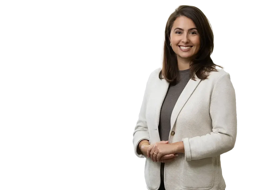 Woman with medium-length brown hair, wearing a white blazer over a dark top, standing and smiling against a plain, black background.