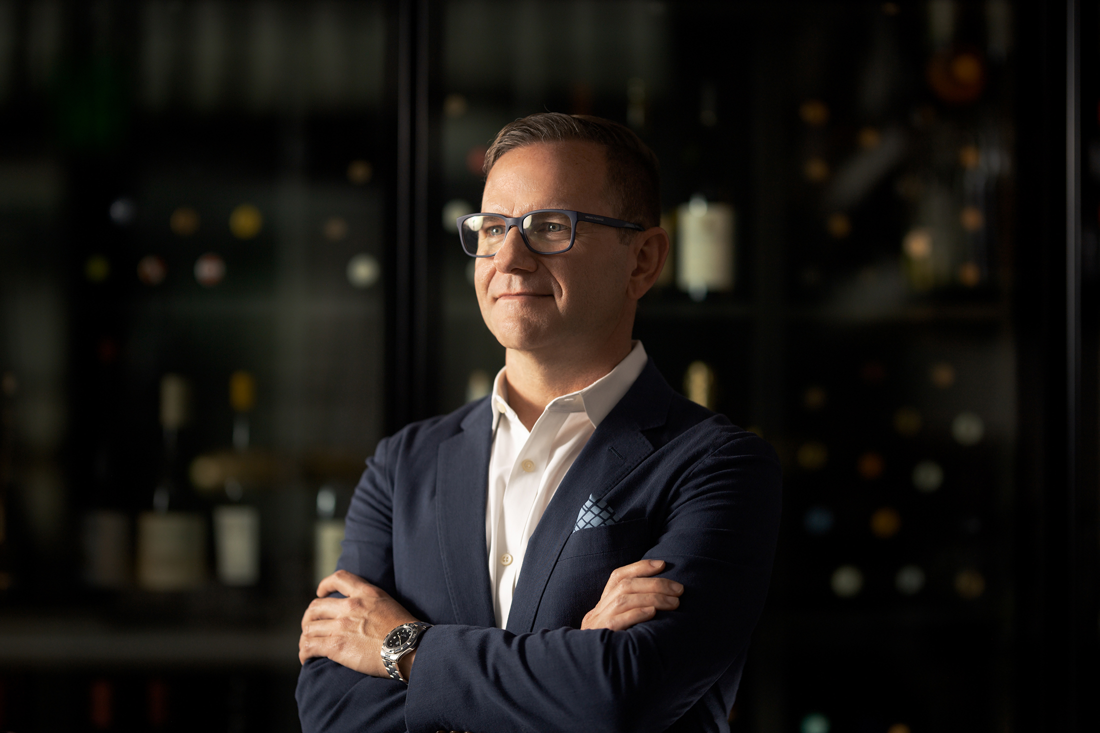 A man in glasses posing creatively among wine bottles for headshot photography.