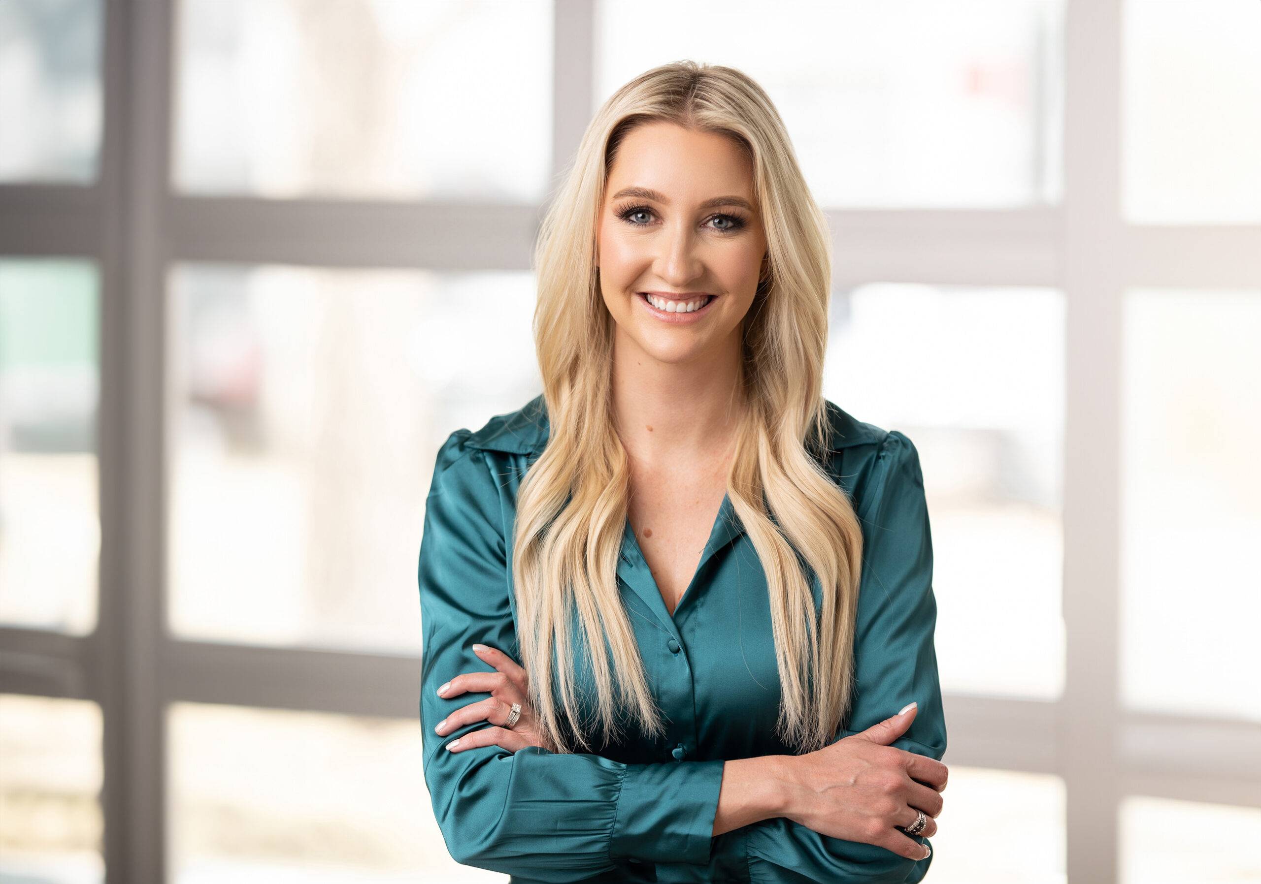 a smiling business woman in a blue shirt standing in front of a window for her headshot for a womens headshot photographer