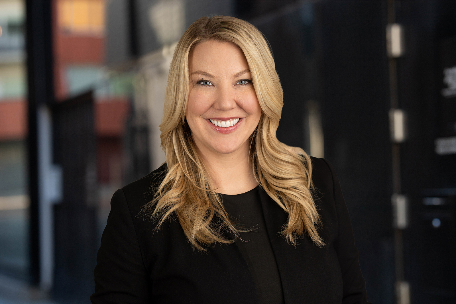 A woman in a black suit, smiling for her headshot against the background of a building.