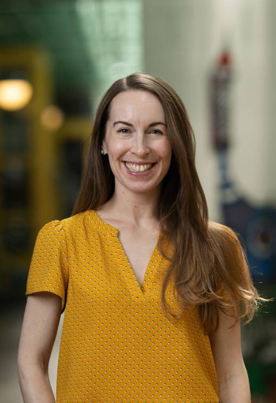 A woman with long brown hair wearing a yellow short-sleeved blouse stands indoors, smiling directly at the camera in this Nimble Team Video.