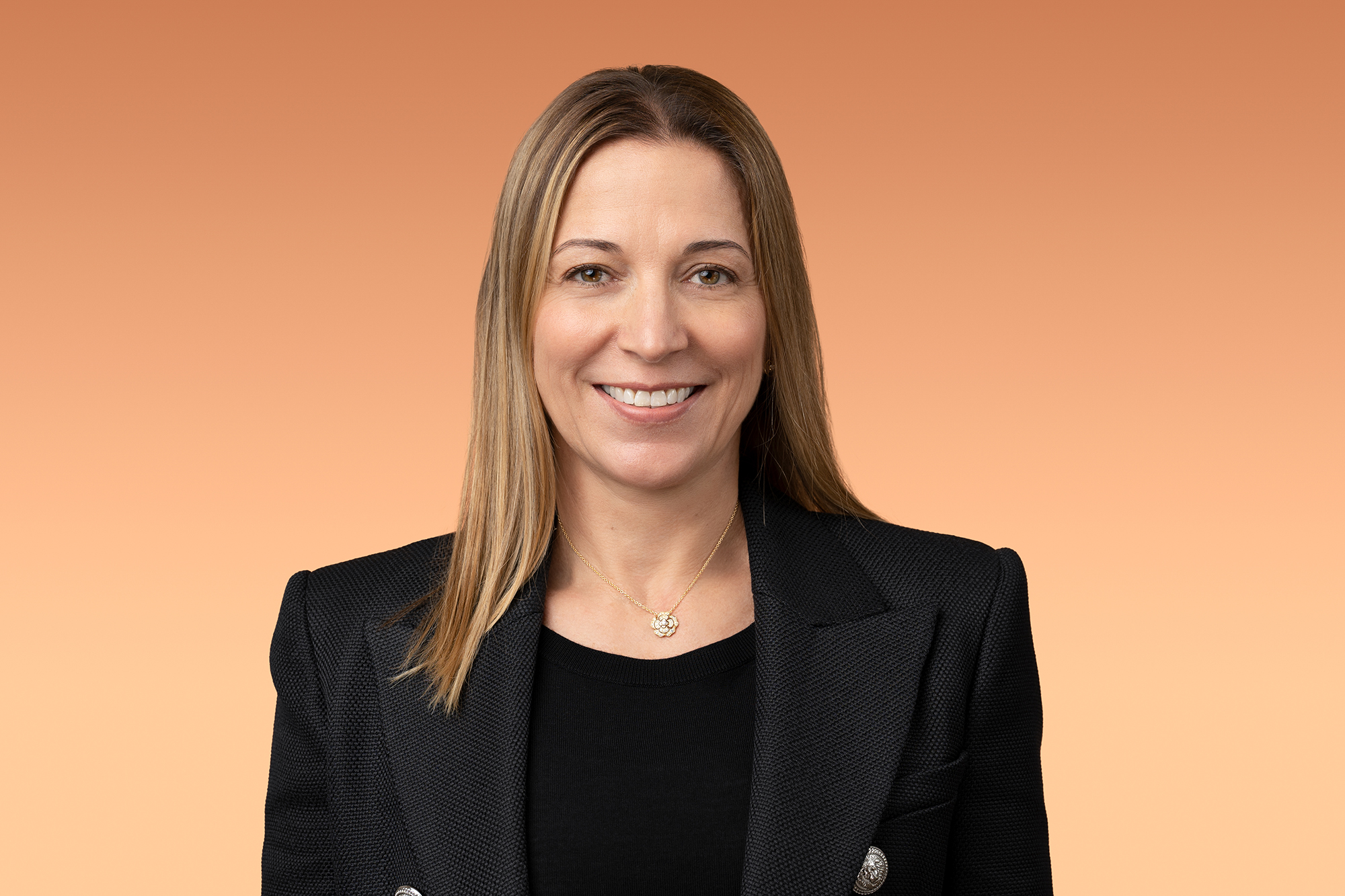A woman in a black blazer posing for a headshot against an orange background.