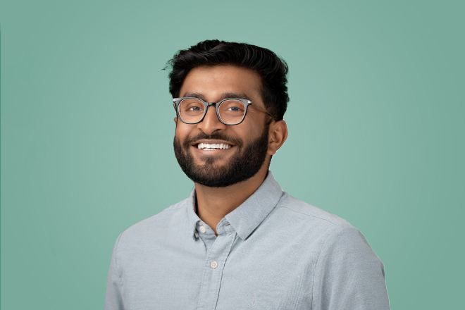 A bearded individual with glasses smiling in front of a green background.