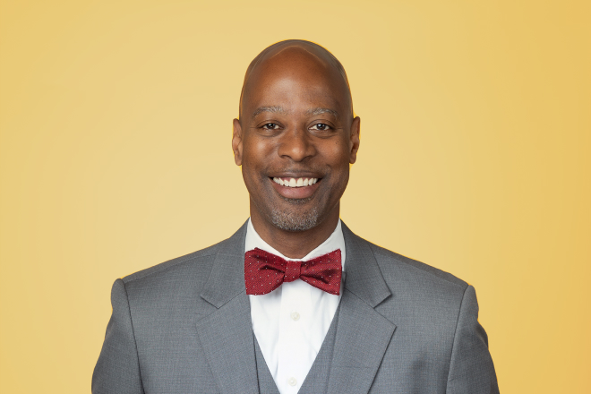 An individual headshot of a black man in a suit and bow tie, smiling against a yellow background.