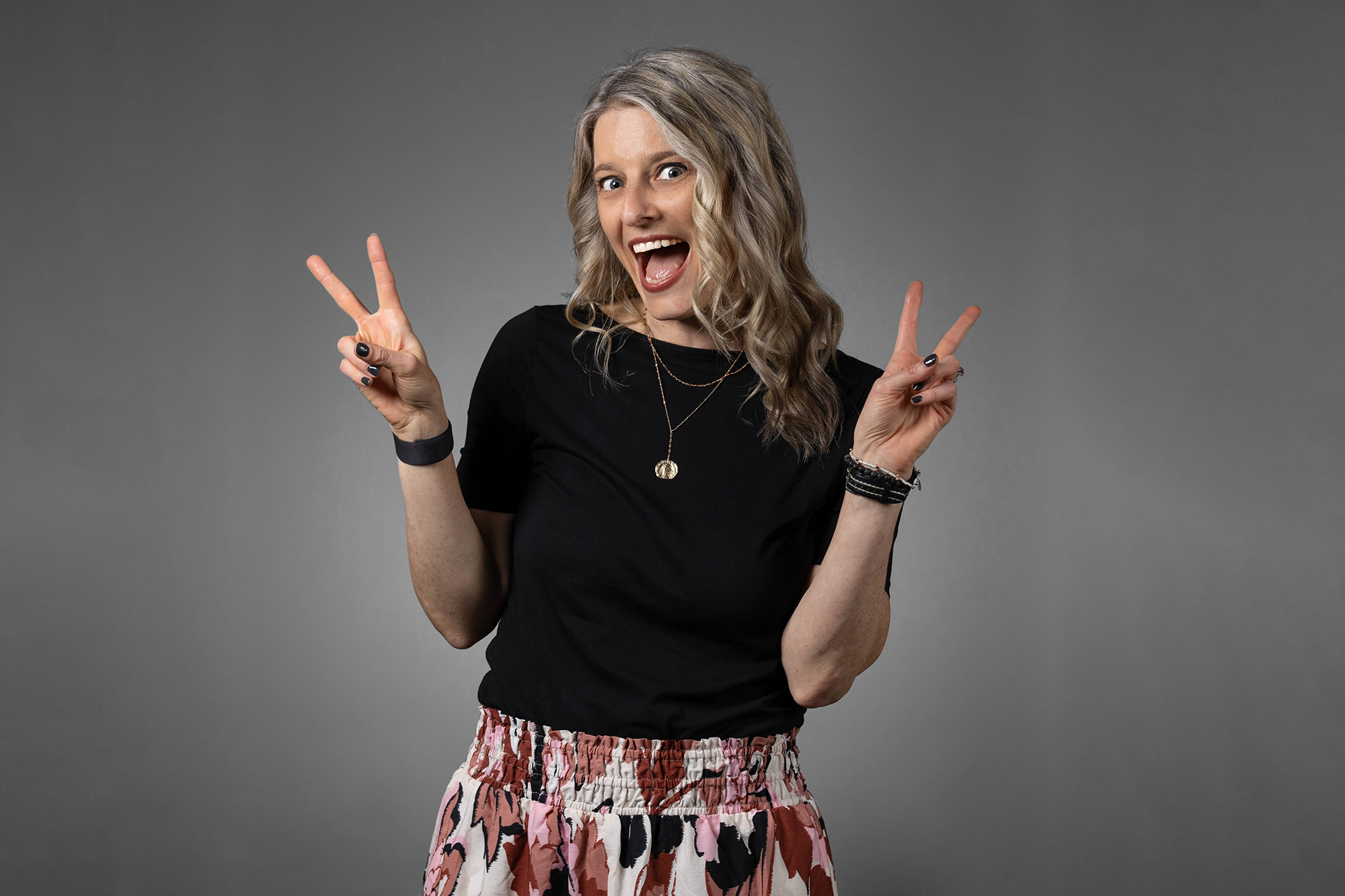 A woman wearing a black shirt and floral skirt making a peace sign poses for corporate headshots.
