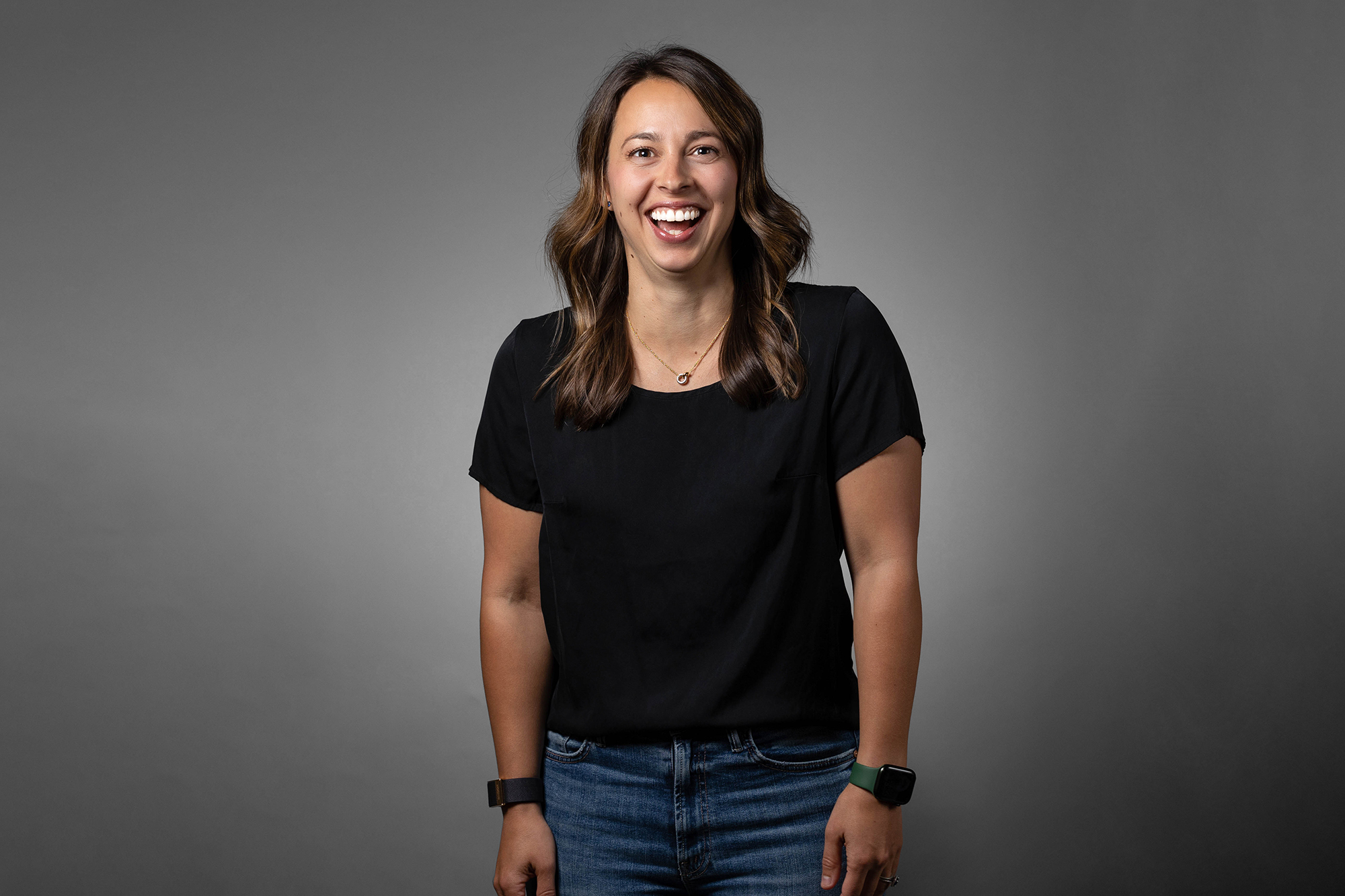 A smiling woman wearing a black shirt and jeans poses for corporate headshots.