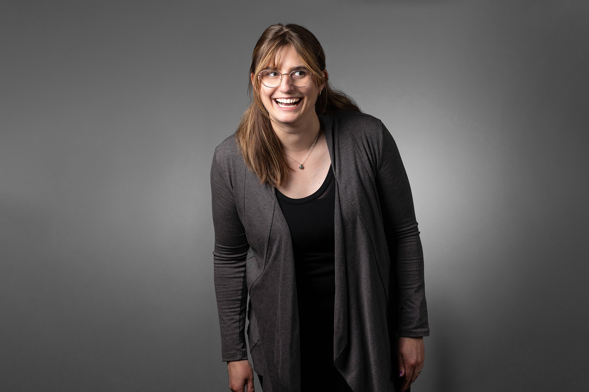 A woman in a gray cardigan is smiling in front of a gray background, presenting her professional corporate headshot.