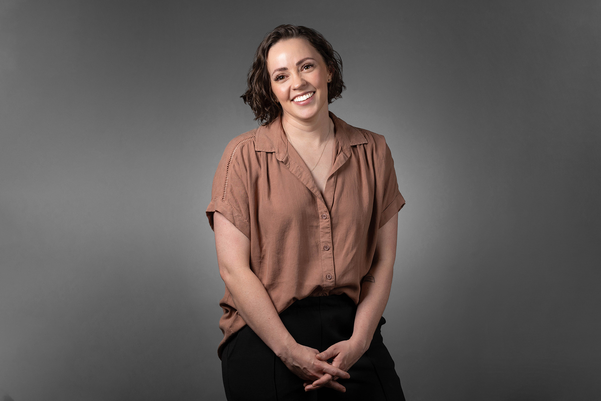 A smiling woman in a brown shirt and black pants poses for corporate headshots.