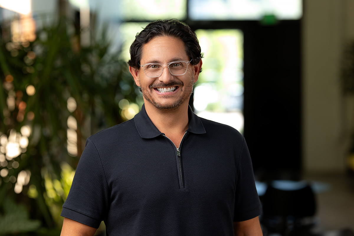 A professional man in a black shirt standing in a hallway, exuding confidence with a friendly smile.