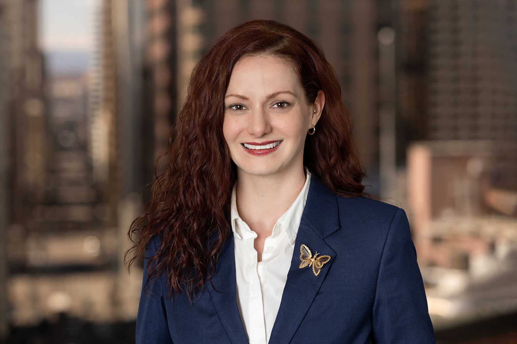 Professional woman with red hair and a navy blazer smiling against a cityscape background in her Denver headshots.