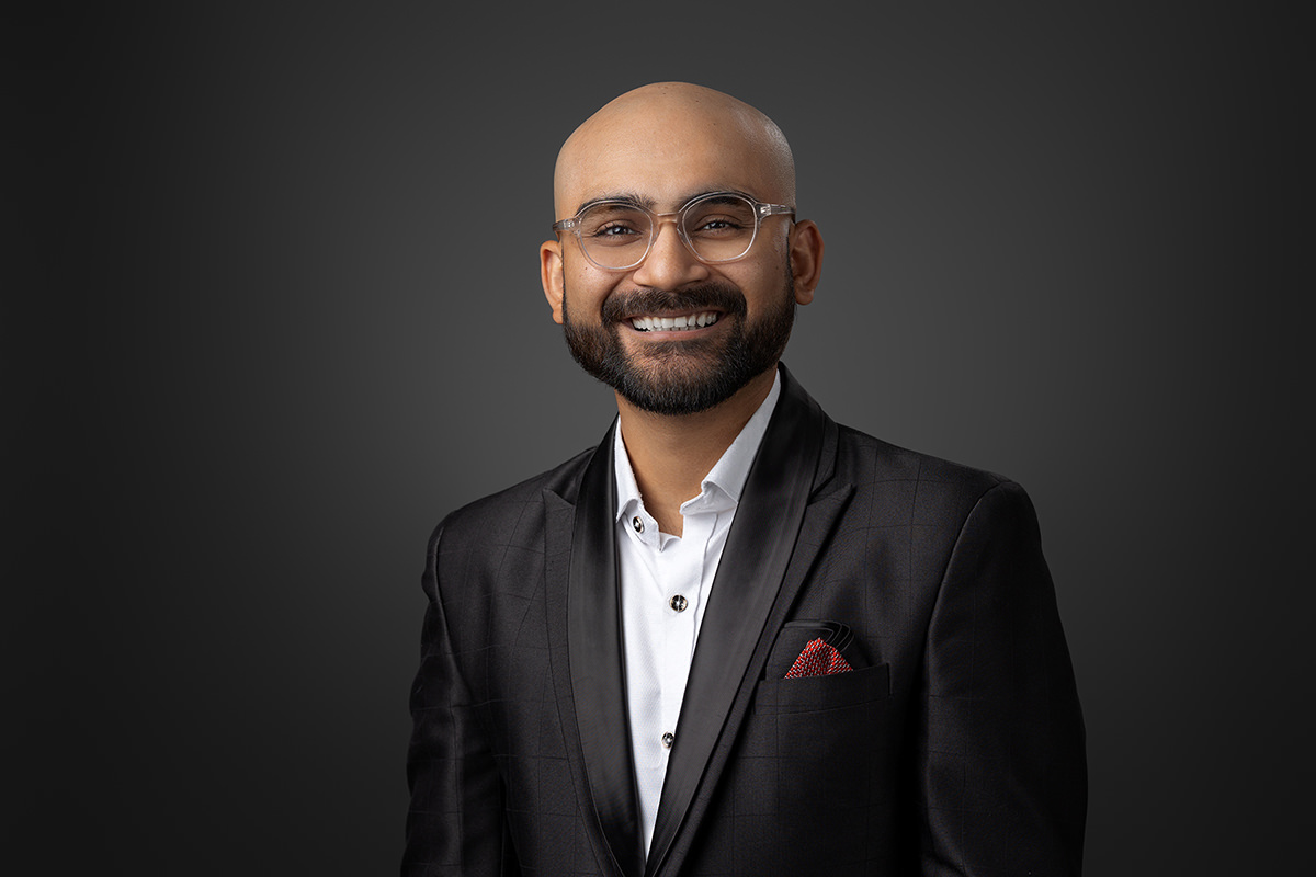 A professional headshot of a smiling man with a beard, wearing glasses, a dark suit, and a white shirt against a gray background.