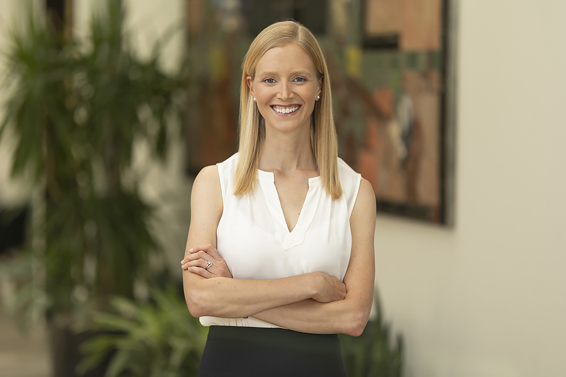 A smiling woman with blonde hair, wearing a white top and dark skirt, stands with her arms crossed in a well-lit indoor setting with paintings in the background, perfect for Denver headshots.