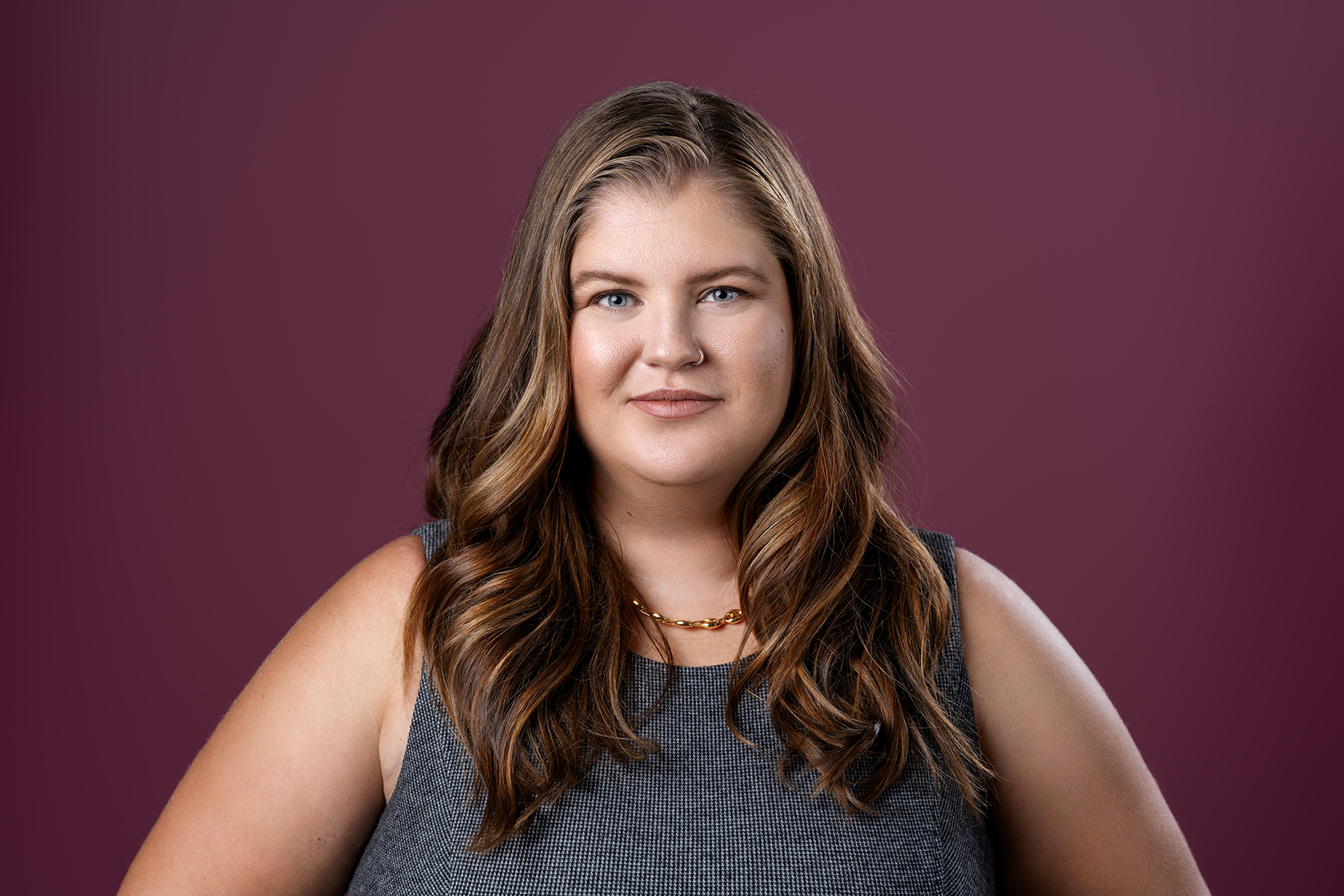 A woman with long wavy hair and a sleeveless top smiles slightly against a purple background. This is an example of Denver headshots.