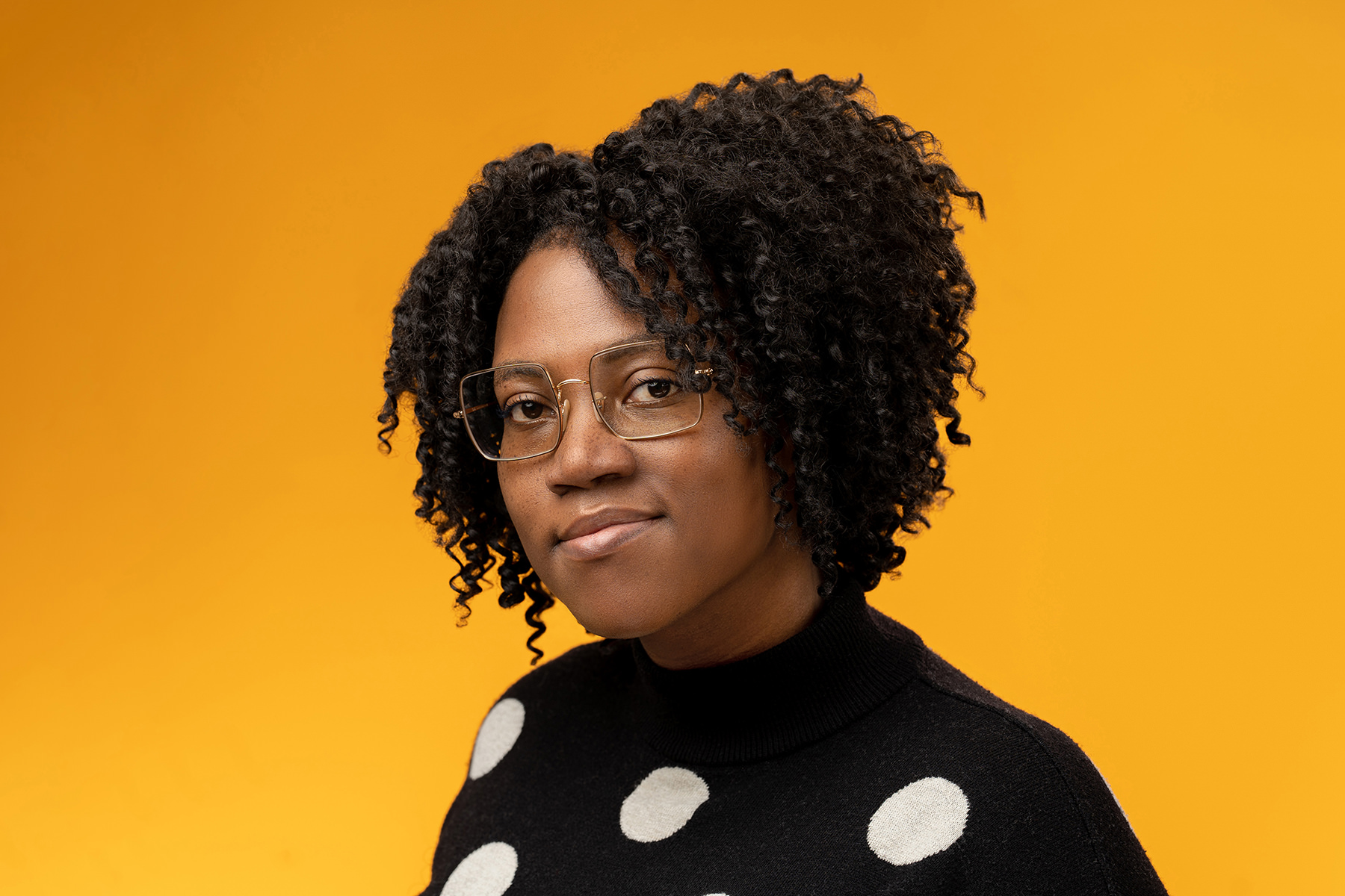 A woman with curly hair wearing glasses and a polka dot sweater poses for her Denver headshots against a yellow background.
