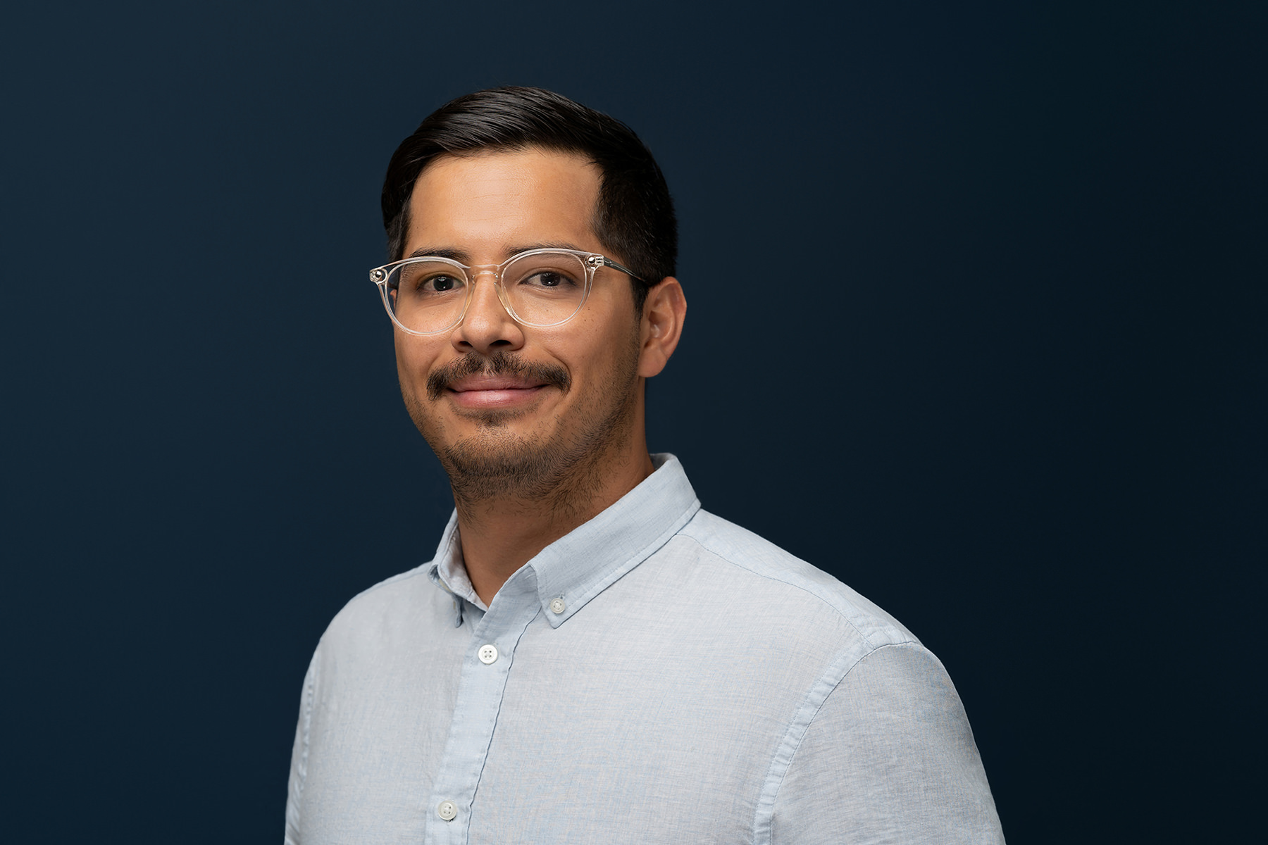 Man with glasses wearing a light blue shirt smiling against a dark blue background in his Denver headshots.