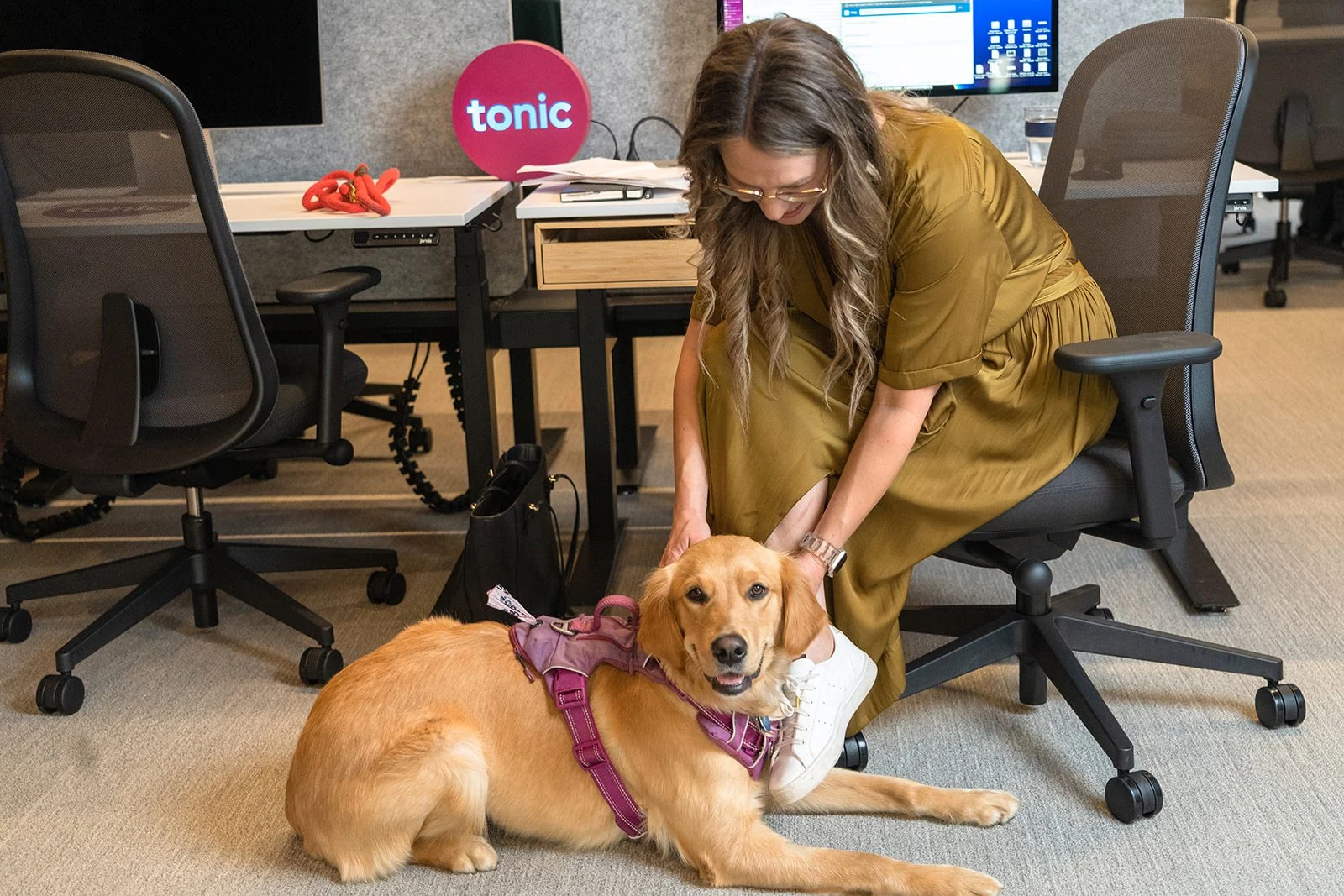 A woman in a green dress pets a golden retriever wearing a harness in an office environment for Denver headshots.