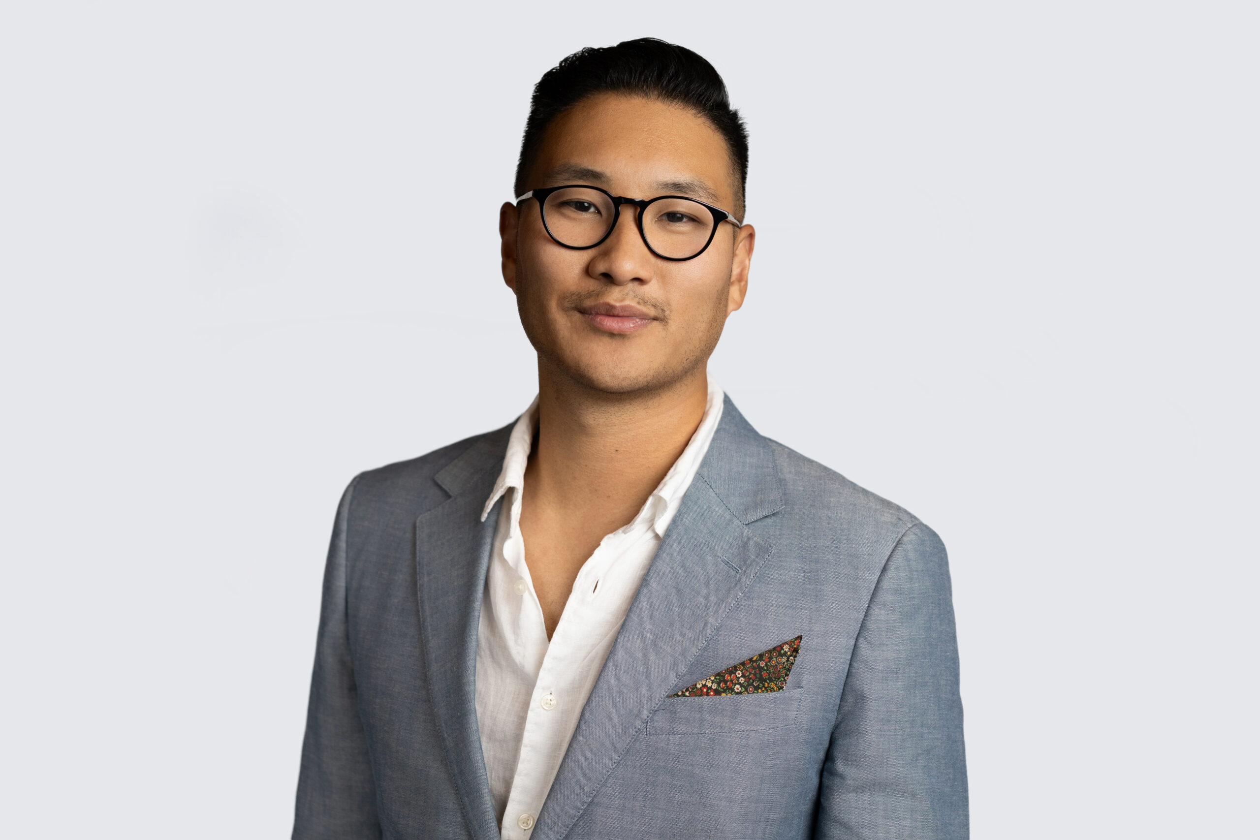 A person wearing glasses and a light gray blazer with a patterned pocket square stands against a plain background, exuding a professional aura perfect for a portfolio headshot.