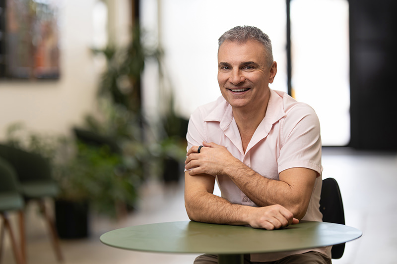 In a bright room adorned with plants, an individual sits at a table, smiling warmly for their headshot.