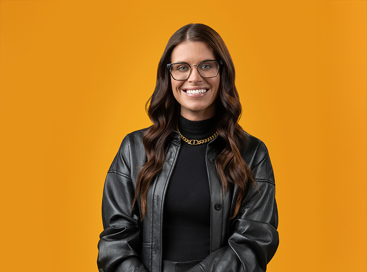 A headshot of a woman with long brown hair, wearing glasses, a black leather jacket, and a gold chain necklace, stands smiling against an orange background.