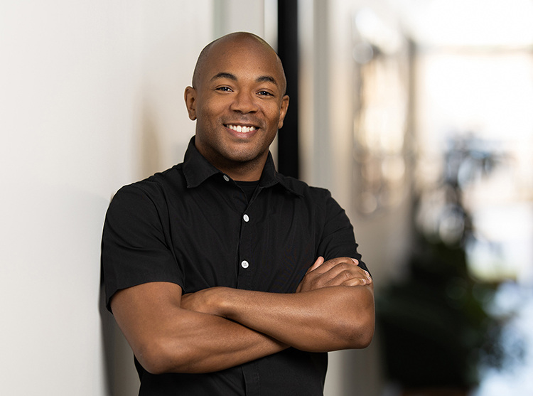 A man wearing a black short-sleeve shirt stands indoors with arms crossed, smiling at the camera in this professional headshot. The background is softly blurred.