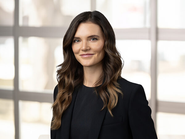 A woman with long brown hair wearing a black blazer smiles slightly in this professional headshot, posing in front of a window with a blurred background.