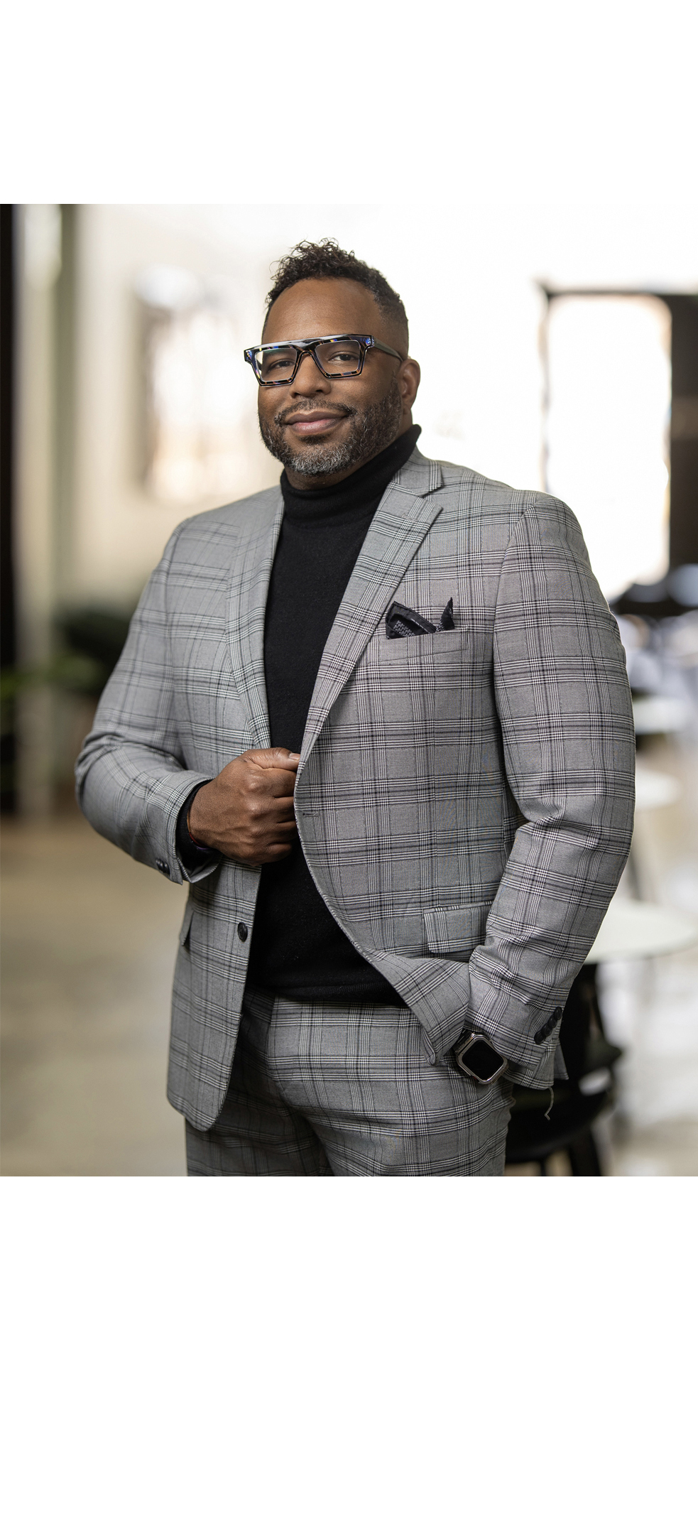 A man in a gray plaid suit and black turtleneck stands indoors, smiling at the camera as if posing for a headshot, with one hand adjusting his jacket.