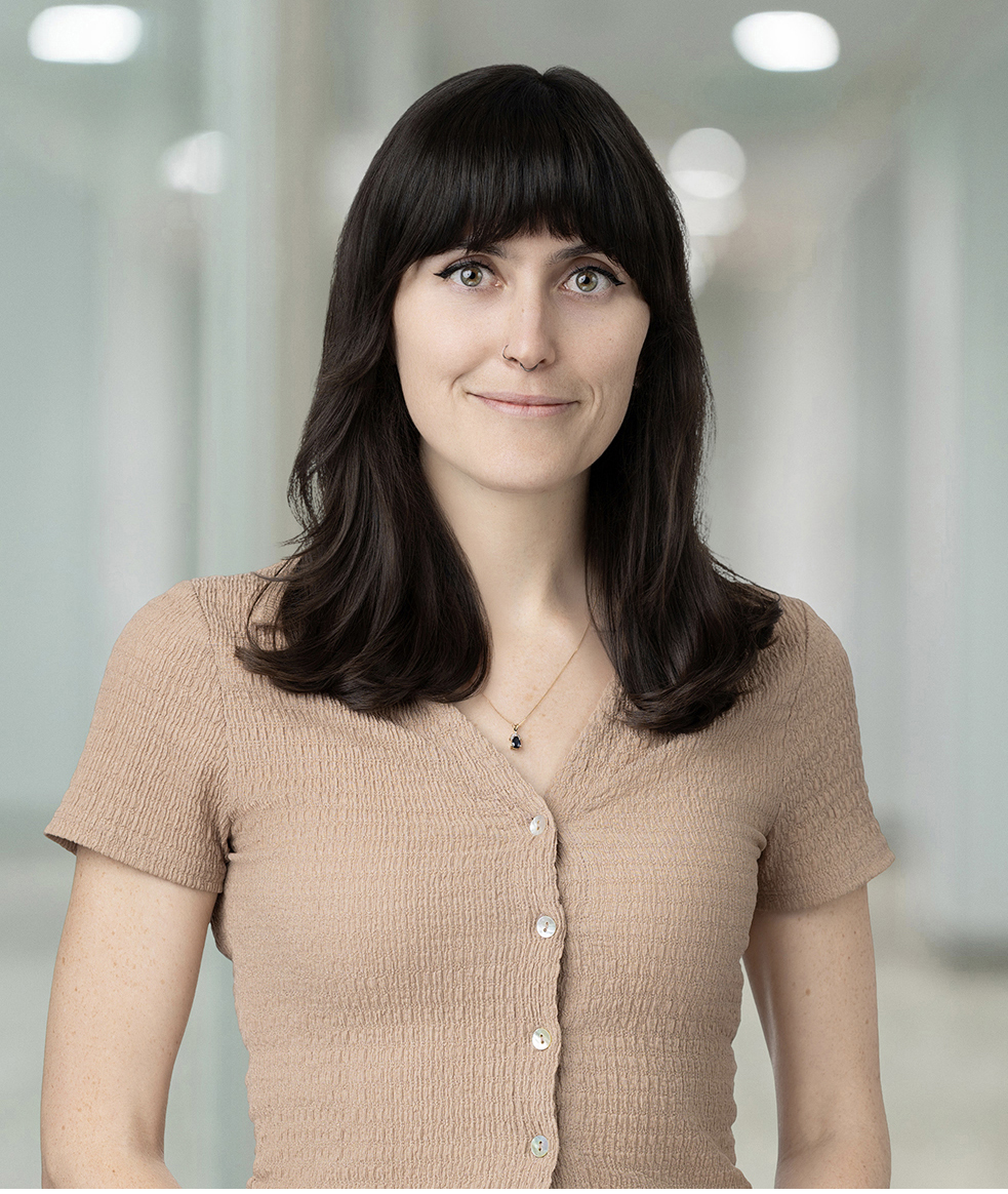 A woman with long dark hair and bangs, wearing a beige short-sleeve button-up top, poses for a headshot indoors against a blurred office background.