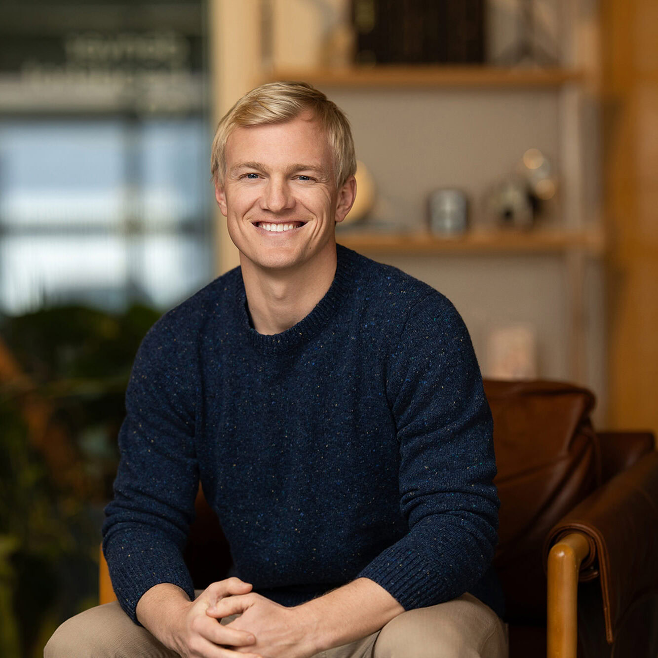 A person with short blond hair, wearing a dark sweater, is seated and smiling in a room adorned with a plant and shelves. This glimpse into their authentic space captures the power of being real amidst the digital world's influence.