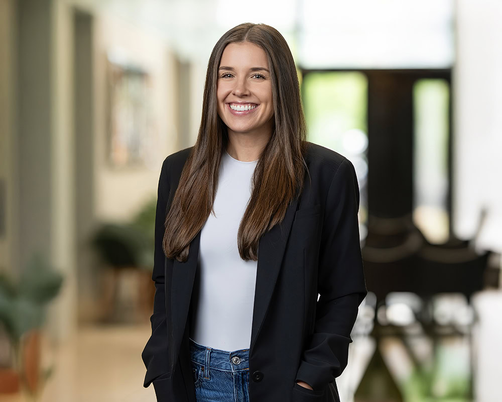 A woman with long brown hair, wearing a black blazer, white top, and jeans, stands indoors with hands in her pockets, smiling at the camera—an example of relaxed individual headshots against a softly blurred background.