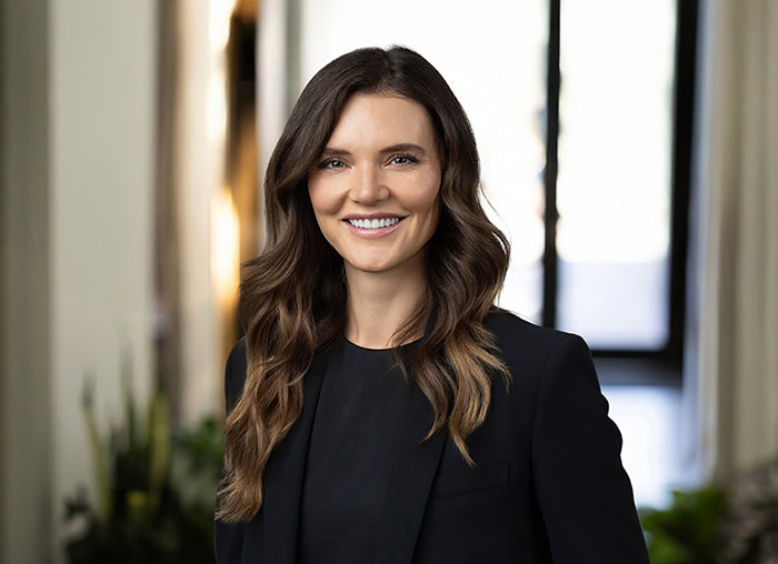 A woman with long brown hair wearing a black blazer and top smiles while standing indoors near a window with plants in the background, perfect for womens headshot photography.