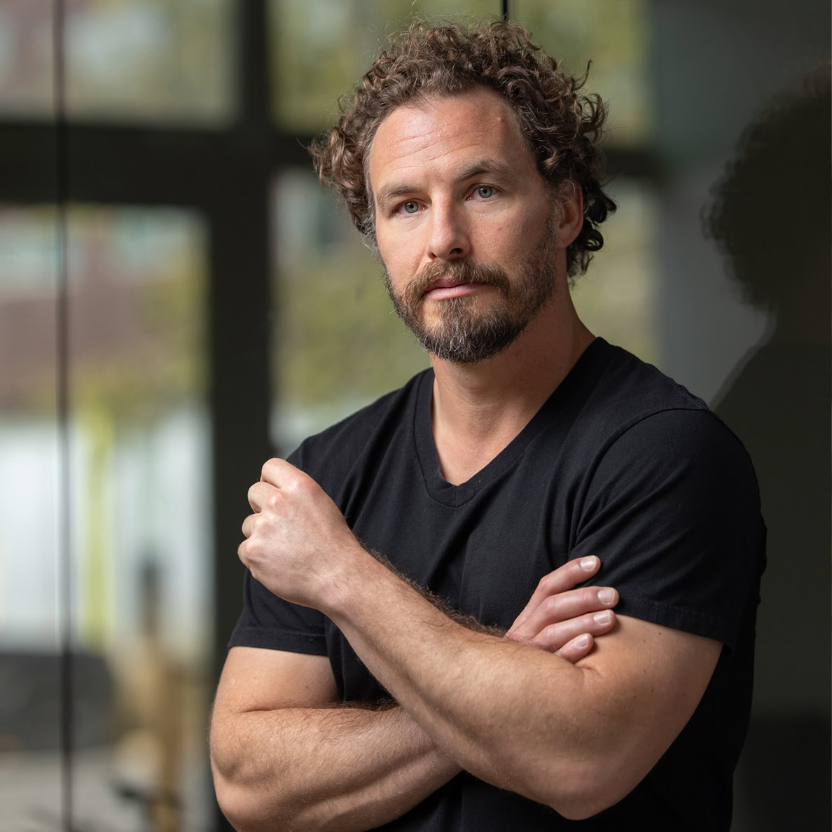 A headshot of a man with curly hair and a beard, wearing a black t-shirt, stands indoors with his arms crossed in front of a window.