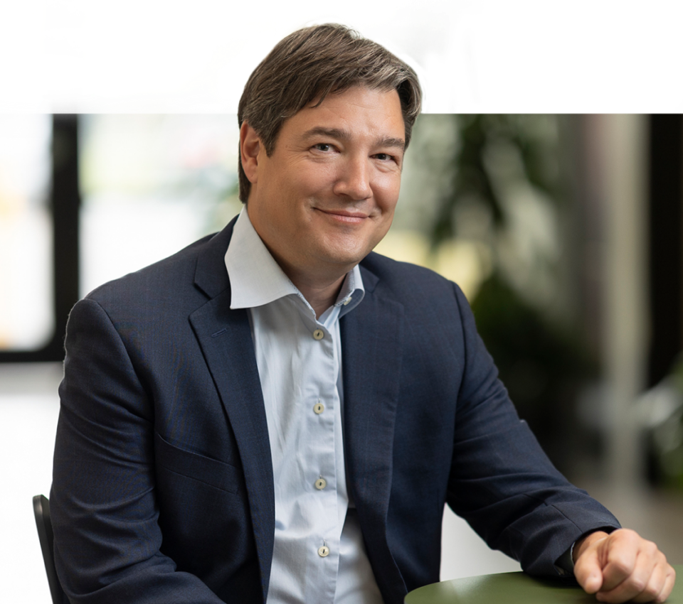 A man in a navy suit jacket and light blue shirt sits at a table, smiling, with a blurred indoor background—perfect for modern headshots or headshots for professionals.
