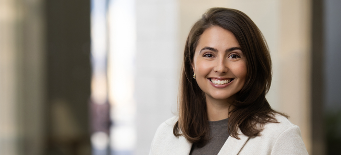A woman with long brown hair, wearing a light blazer and grey top, smiles at the camera in a bright indoor setting—perfect for modern headshots.