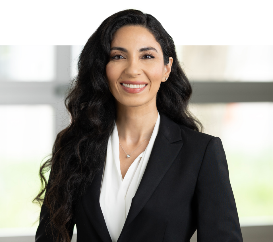 A woman with long dark hair wearing a black blazer and white blouse smiles at the camera in a bright indoor setting, perfect for modern headshots for professionals.