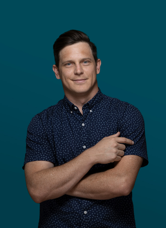 Man with short dark hair wearing a navy patterned short-sleeve button-up shirt, standing against a solid teal background and pointing with his right hand—perfect for modern headshots or headshots for professionals.